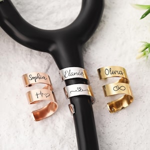 Puede incluir: Tres brazaletes metálicos en espiral para estetoscopio, con acabados en oro rosa, plata y oro. Cada brazalete está grabado con un nombre, incluyendo Sophia, Elanie y Olivia, junto a símbolos como un latido, un corazón y el signo de infinito.