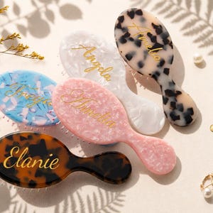 May include: Four oval hairbrushes with handles, each featuring a unique marbled pattern in blue, pink, white, or brown. Each brush is personalized with a gold cursive name: Freya, Amelia, Angela, and Elanie, printed on the smooth surface.