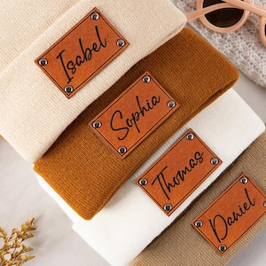 May include: Four folded knit beanies in cream, mustard yellow, white, and tan colors. Each beanie features a rectangular brown faux leather patch attached with four silver metal rivets. The patches are engraved with the names Isabel, Sophia, Thomas, and Daniel in a black cursive script font.