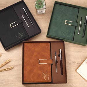 May include: Three gift sets each containing a rectangular notebook with a gold-toned metal clasp, a matching ballpoint pen, and a refill cartridge. The sets come in black, dark green, and brown, with personalised letter monograms on the covers.