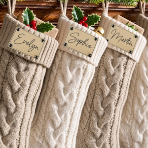 May include: Three cream-colored cable knit Christmas stockings featuring rectangular gold-toned metal nameplates engraved with the names Evelyn, Sophia, and Martin. Each stocking is adorned with a sprig of green holly and red berries.