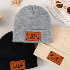 Op de afbeelding: Drie gebreide mutsen in grijs, zwart en beige, elk voorzien van een rechthoekige bruine, leerachtige patch die aan de voorzijde is vastgeklonken. De patches zijn voorzien van de namen Charles, Rhoda en Michael in een eenvoudig, donker lettertype.