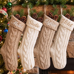 Puede incluir: Cuatro calcetines navideños de punto trenzado en colores beige y crema alternos, cada uno con un puño doblado bordado con los nombres Mateo, Sadie, Dylan y Violet. Cada calcetín está relleno de agujas de pino verdes y piñas marrones.