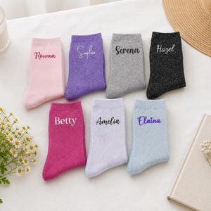 May include: Seven pairs of socks in various colours including pink, purple, grey, black, magenta, light purple, and light blue. Each sock features a unique name embroidered in a script or print font: Rowan, Sophia, Serena, Hazel, Betty, Amelia, and Elaina.