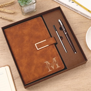 May include: A brown leather-effect notebook with a gold-coloured metal clasp and the name MICHAEL engraved on the cover, presented in a gift box alongside a matching brown pen and a silver-coloured ink refill, all arranged in a structured display set.