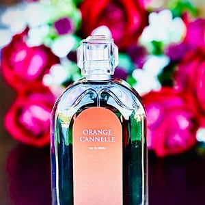 Könnte beinhalten: Ein klarer Glasparfümflakon mit einem rechteckigen orangefarbenen Etikett mit der Aufschrift "ORANGE CANNELLE eau de toilette". Die Flasche hat einen transparenten, geometrischen Verschluss. Der Hintergrund zeigt ein verschwommenes Arrangement aus rosa, roten und weißen Blumen.