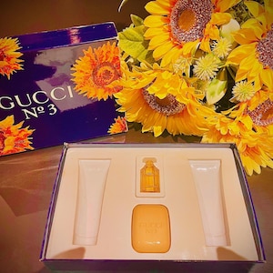 Puede incluir: Un juego de regalo Gucci No. 3 vintage con una caja con ilustraciones de girasoles. El juego incluye un pequeño frasco de perfume, una pastilla de jabón y dos tubos de loción. Un ramo de girasoles está en el fondo.