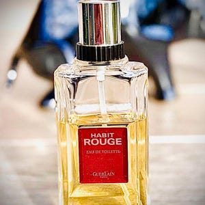 May include: A clear glass bottle of Habit Rouge Eau de Toilette perfume. The bottle has a silver cap and a red label with white text. The liquid inside is amber-coloured. The background is blurred.