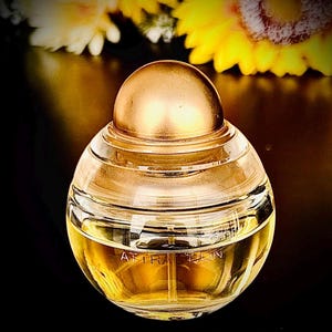 Op de afbeelding: Een ronde, heldere glazen parfumfles met een goudkleurige dop en accenten. De fles is gevuld met een gouden vloeistof en het woord "ATTRACTION" staat op de voorkant. De achtergrond toont gele en witte bloemen.