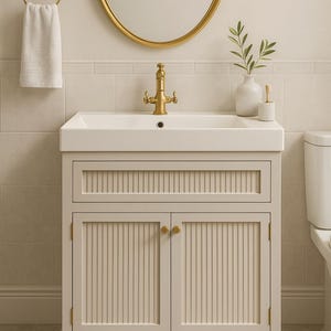 May include: A cream-coloured bathroom vanity unit with a white rectangular sink and gold-coloured tap. The cabinet has vertical paneling on the doors and drawer. A gold-framed mirror and towel ring are also visible.
