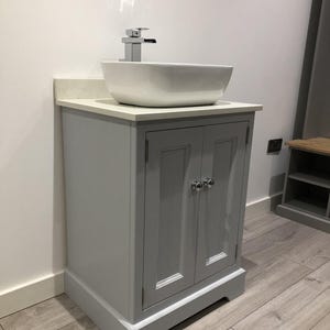 May include: A grey bathroom vanity unit with a white countertop and a white, rectangular vessel sink. The vanity has two doors with silver knobs and a chrome tap. The base of the unit is a darker shade of grey.