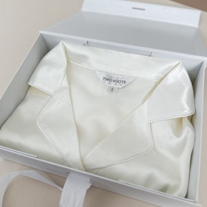 Puede incluir: Una camisa de seda blanca con cuello, doblada y colocada dentro de una caja de regalo blanca con una cinta blanca. La camisa tiene una etiqueta que dice "TWO KNOTS" y "S".