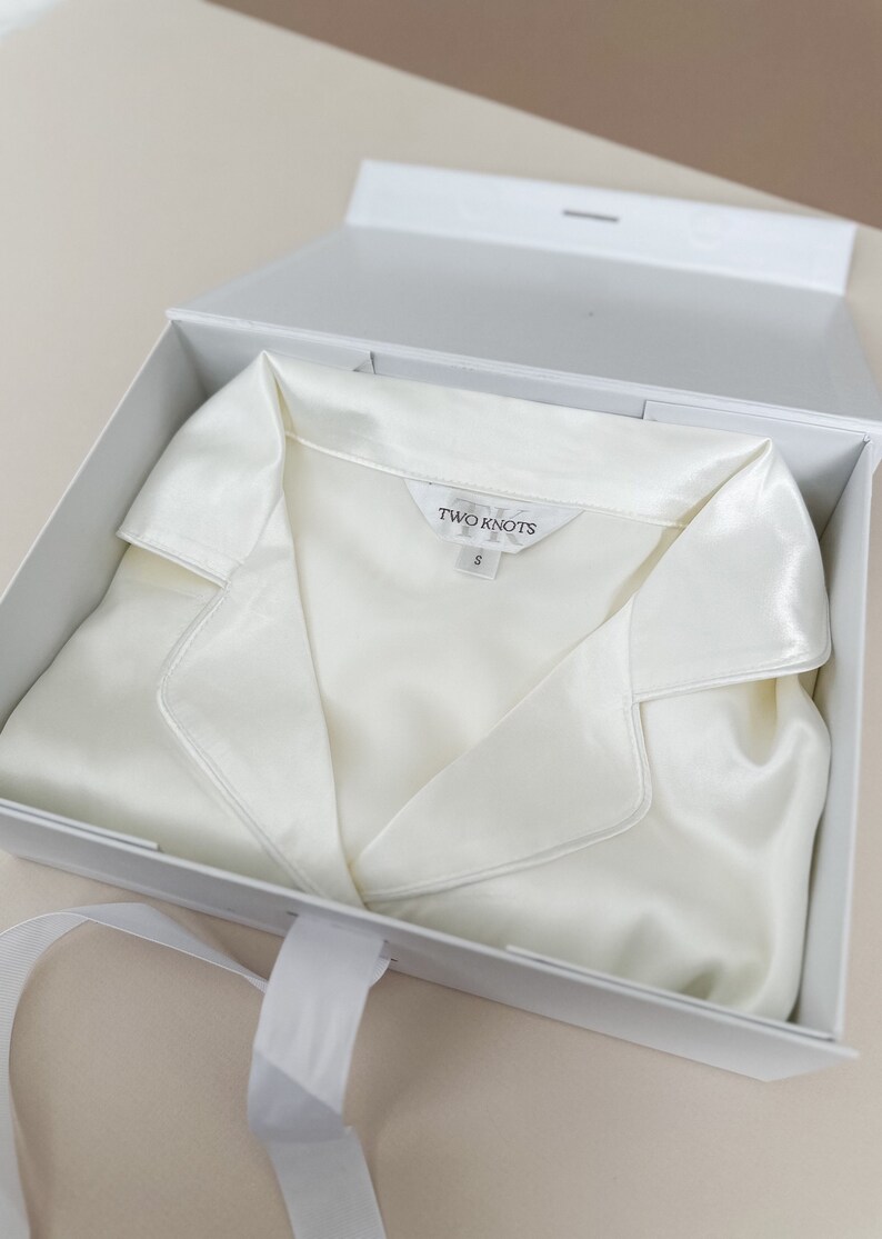 Puede incluir: Una camisa de seda blanca con cuello, doblada y colocada dentro de una caja de regalo blanca con una cinta blanca. La camisa tiene una etiqueta que dice "TWO KNOTS" y "S".