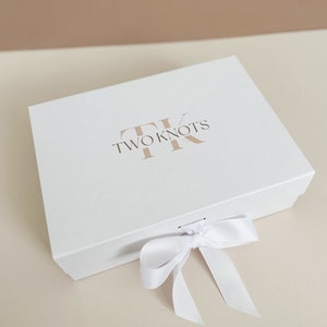 Puede incluir: Una caja de regalo blanca con una cinta de sat&eacute;n blanca atada en un lazo. La caja tiene el texto "TWO KNOTS" y un logotipo "TK" estilizado impreso en la parte superior.