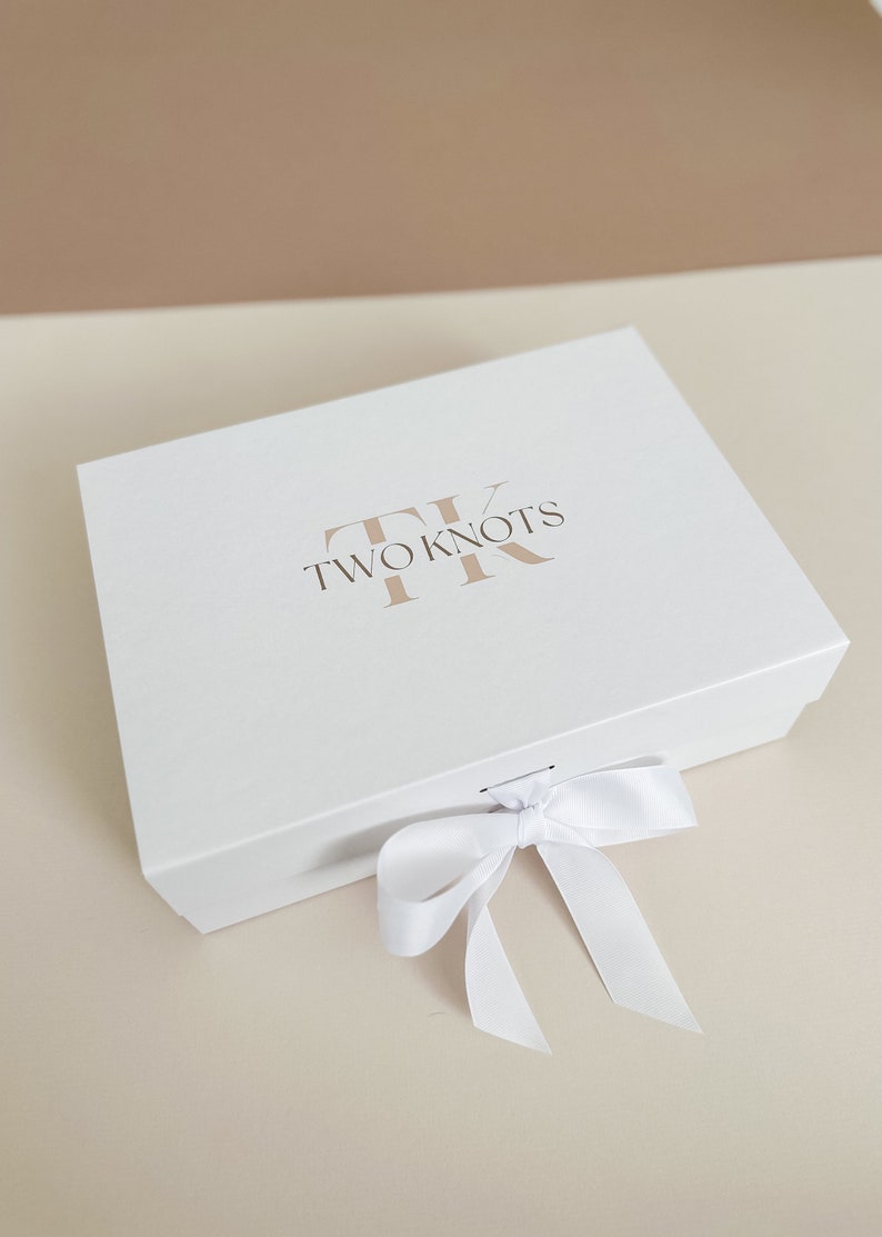 Puede incluir: Una caja de regalo blanca con una cinta de sat&eacute;n blanca atada en un lazo. La caja tiene el texto "TWO KNOTS" y un logotipo "TK" estilizado impreso en la parte superior.