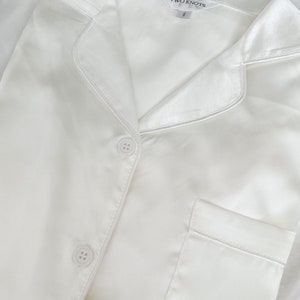 Puede incluir: Camiseta de pijama de sat&eacute;n blanco con cierre de botones y un bolsillo. La camiseta tiene cuello y la etiqueta dice "TWO KNOTS" con la talla "S".