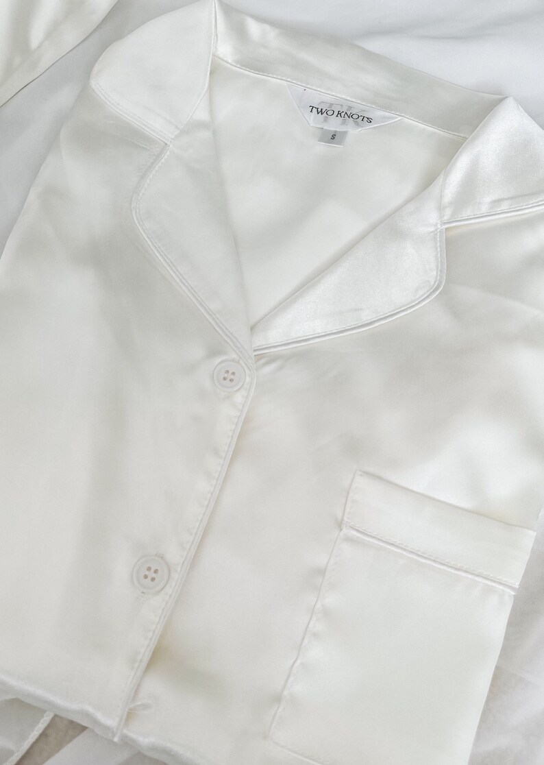 Puede incluir: Camiseta de pijama de sat&eacute;n blanco con cierre de botones y un bolsillo. La camiseta tiene cuello y la etiqueta dice "TWO KNOTS" con la talla "S".