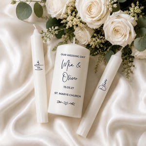 Op de afbeelding: Drie witte waskaarsen, één dikke stompkaars en twee slanke dinerkaarsen. De stompkaars bevat de tekst "OUR WEDDING DAY", "Mia & Oliver", "15.05.27", "ST. MARYS CHURCH" en een hartje. De dinerkaarsen zijn bedrukt met "Mia" en "Oliver".