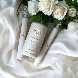 May include: A collection of white wedding candles, decorated with floral designs and text. The central pillar candle features a floral motif and the inscription "THE WEDDING OF Steve & Mel 20.06.25 St Marys Church". Two taper candles are also present.