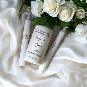 May include: A set of white candles, including a pillar candle and two tapers, with black calligraphy. The pillar candle reads "Welcome to our Happily Ever After Mia & Oliver 15.05.25 St. Marys Church". White roses and foliage are arranged in the background.