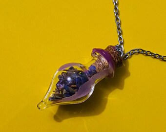 Peut inclure: Un collier en chaîne argentée avec un pendentif en forme de bouteille en verre. La bouteille est remplie de fleurs séchées violettes et possède un bouchon en liège avec une bande violette.