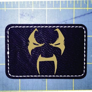 May include: A rectangular patch with a dark blue background and a gold-coloured graphic of a stylised face. The patch is outlined with white stitching.