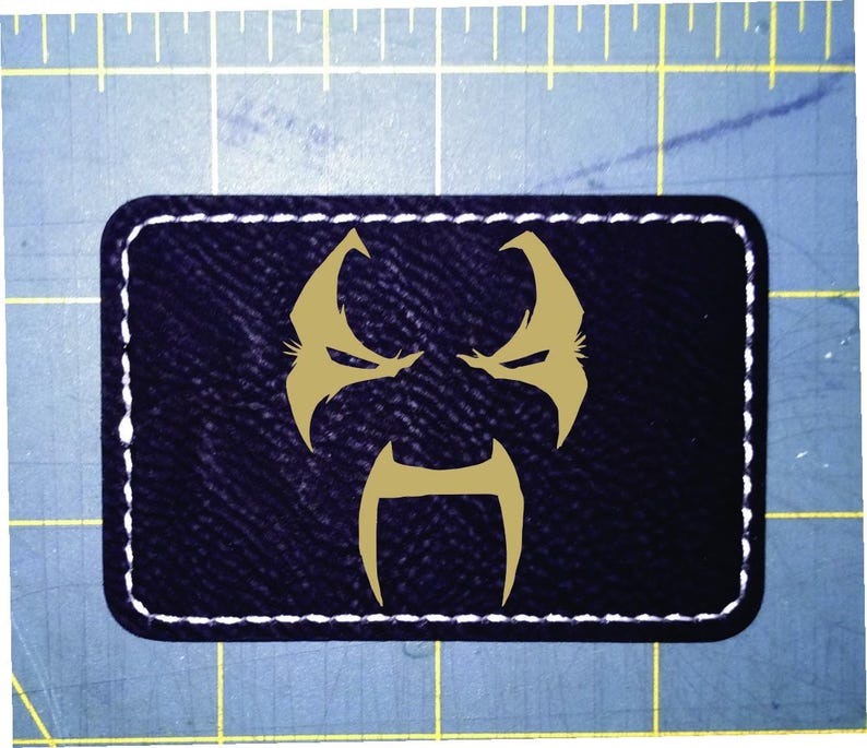 May include: A rectangular patch with a dark blue background and a gold-coloured graphic of a stylised face. The patch is outlined with white stitching.