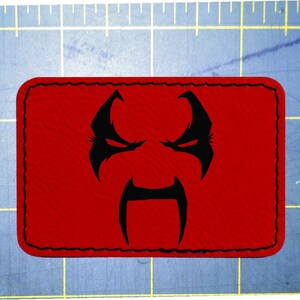 Op de afbeelding: Een rechthoekige rode patch met een zwart gestikte rand. De patch heeft een zwarte afbeelding van een gestileerd gezicht met een boze uitdrukking. Het gezicht heeft puntige wenkbrauwen, ogen en een lange, gebogen snor.