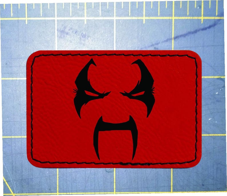 May include: A rectangular red patch with a black stitched border. The patch features a black graphic of a stylized face with an angry expression. The face has pointed eyebrows, eyes, and a long, curved moustache.