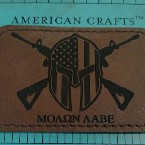 Op de afbeelding: Een rechthoekige bruine leren patch met een zwarte afbeelding van een Spartaanse helm, gekruiste geweren en de Griekse zin "MOLON LABE". De patch is rondom gestikt en heeft de tekst "AMERICAN CRAFTS" bovenaan.