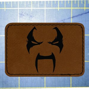 May include: A brown rectangular patch with a black stitched border. The patch features a black graphic of a stylized face with an angry expression, including eyebrows, eyes, and a mustache-like shape.