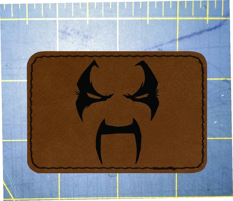 May include: A brown rectangular patch with a black stitched border. The patch features a black graphic of a stylized face with an angry expression, including eyebrows, eyes, and a mustache-like shape.