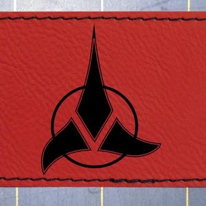 Può includere: Una toppa in pelle rossa con bordo cucito nero. La toppa presenta un emblema nero con un design circolare e una forma a tre punte. La toppa è rettangolare con angoli arrotondati.