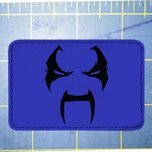 May include: A rectangular blue patch featuring a black graphic of a stylized face. The face has a stern expression with angled eyebrows, eyes, and a long, curved moustache. The patch has a stitched border.