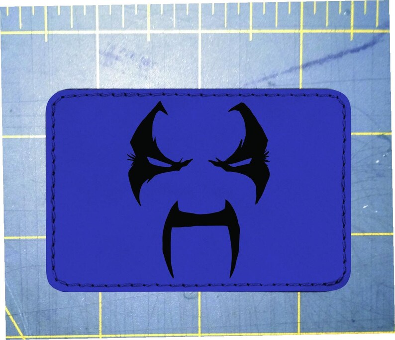 May include: A rectangular blue patch featuring a black graphic of a stylized face. The face has a stern expression with angled eyebrows, eyes, and a long, curved moustache. The patch has a stitched border.