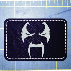 May include: A rectangular black patch with a white stitched border. The patch features a stylized, light grey face design. The design includes two angular shapes for eyes and a central, inverted U-shape.