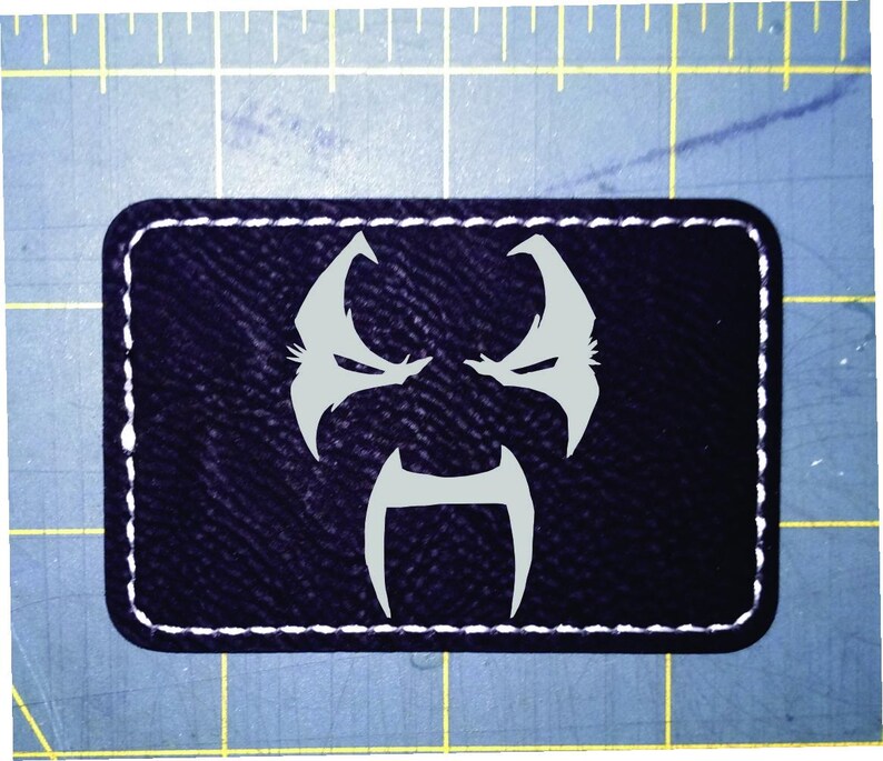 May include: A rectangular black patch with a white stitched border. The patch features a stylized, light grey face design. The design includes two angular shapes for eyes and a central, inverted U-shape.