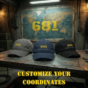 Puede incluir: Tres gorras de béisbol en gris, azul marino y negro, cada una con el texto amarillo 681 y coordenadas geográficas bordadas en la parte frontal. Las gorras tienen un borde desgastado y se muestran sobre una superficie metálica ante un mapa.