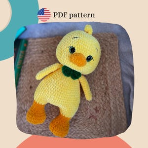 May include: A handmade yellow crochet duck with orange feet and beak, and a green bow tie. The duck has black eyes and is labeled "PDF pattern". The plush toy is on a textured brown surface.