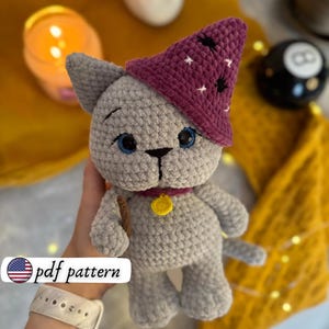 May include: A grey crocheted cat doll wearing a purple wizard's hat with white star details. The cat has blue eyes, a yellow pendant, and a small wand. The text "pdf pattern" is in the lower left corner.