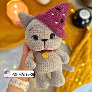 Könnte beinhalten: Eine gehäkelte graue Katze mit einem lila Zaubererhut mit weißen Sternen. Die Katze hat blaue Augen, ein rosa Halsband mit einem goldenen Anhänger und einen kleinen Zauberstab. Der Text "PDF PATTERN" ist enthalten.