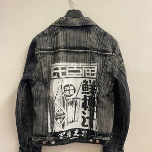 May include: A black denim jacket with a white graphic on the back. The graphic features a bottle of "Wild Strawberry Beverage" and Japanese text. The jacket has a classic denim style with a button closure and two pockets.