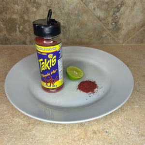 May include: A clear glass bottle of Takis Flamin' Hot seasoning with a black lid, a lime wedge, and a pile of red seasoning on a white plate. The bottle has a blue and yellow label with the Takis logo.