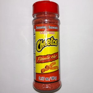 May include: A clear plastic bottle of Cheetos Flamin' Hot seasoning with a red lid. The label is yellow with the Cheetos logo and the words "Flamin' Hot". The bottle contains 90 g of red seasoning.