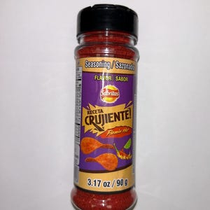 May include: A clear plastic bottle of Sabritas Flamin' Hot seasoning. The label is purple and features the words "RECETA CRUJIENTE!" and "Flamin' Hot." The bottle contains 90 g of red seasoning and has a black lid.
