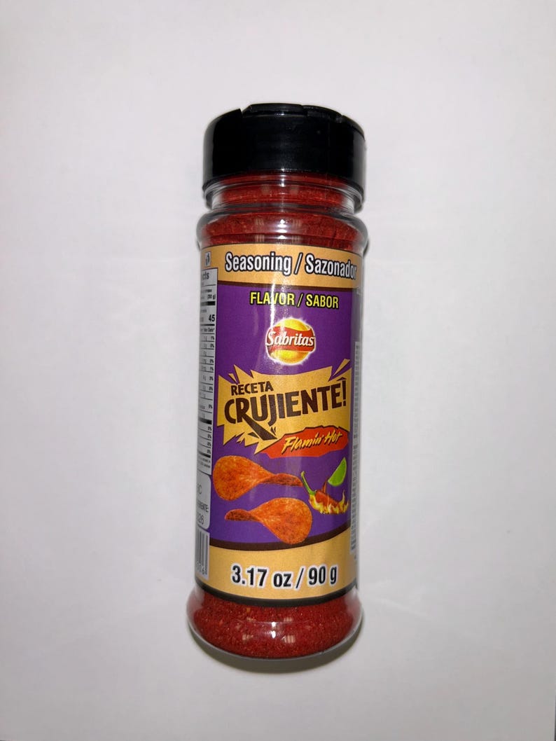 May include: A clear plastic bottle of Sabritas Flamin' Hot seasoning. The label is purple and features the words "RECETA CRUJIENTE!" and "Flamin' Hot." The bottle contains 90 g of red seasoning and has a black lid.