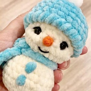 May include: A crocheted snowman toy with a light blue hat and scarf. The snowman has a white body, black eyes, an orange nose, and a smiling mouth. The hat features a white pom-pom. The figure is made of a soft, textured material.