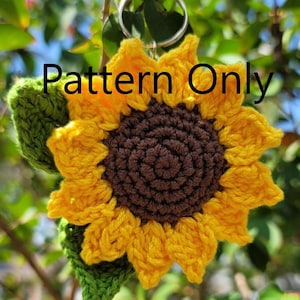 May include: Crocheted sunflower keychain with yellow petals, a brown center, and green leaves. The keychain is attached to a silver ring. The text "Pattern Only" is displayed on the sunflower.