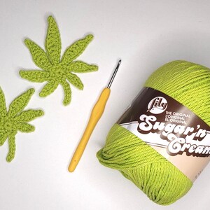 May include: Two crocheted green cannabis leaves, a yellow crochet hook, and a ball of green yarn labeled "Sugar'n Cream" are arranged on a white surface. The yarn is a bright, solid green color, and the leaves are detailed.