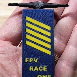 Op de afbeelding: Een rechthoekig blauw 3D-geprint object met een getextureerd oppervlak, voorzien van vijf diagonale gele strepen en de gele tekst FPV RACE ONE eronder. Een zwarte T-vormige antenne is bevestigd aan de bovenkant van de rechthoekige basis.